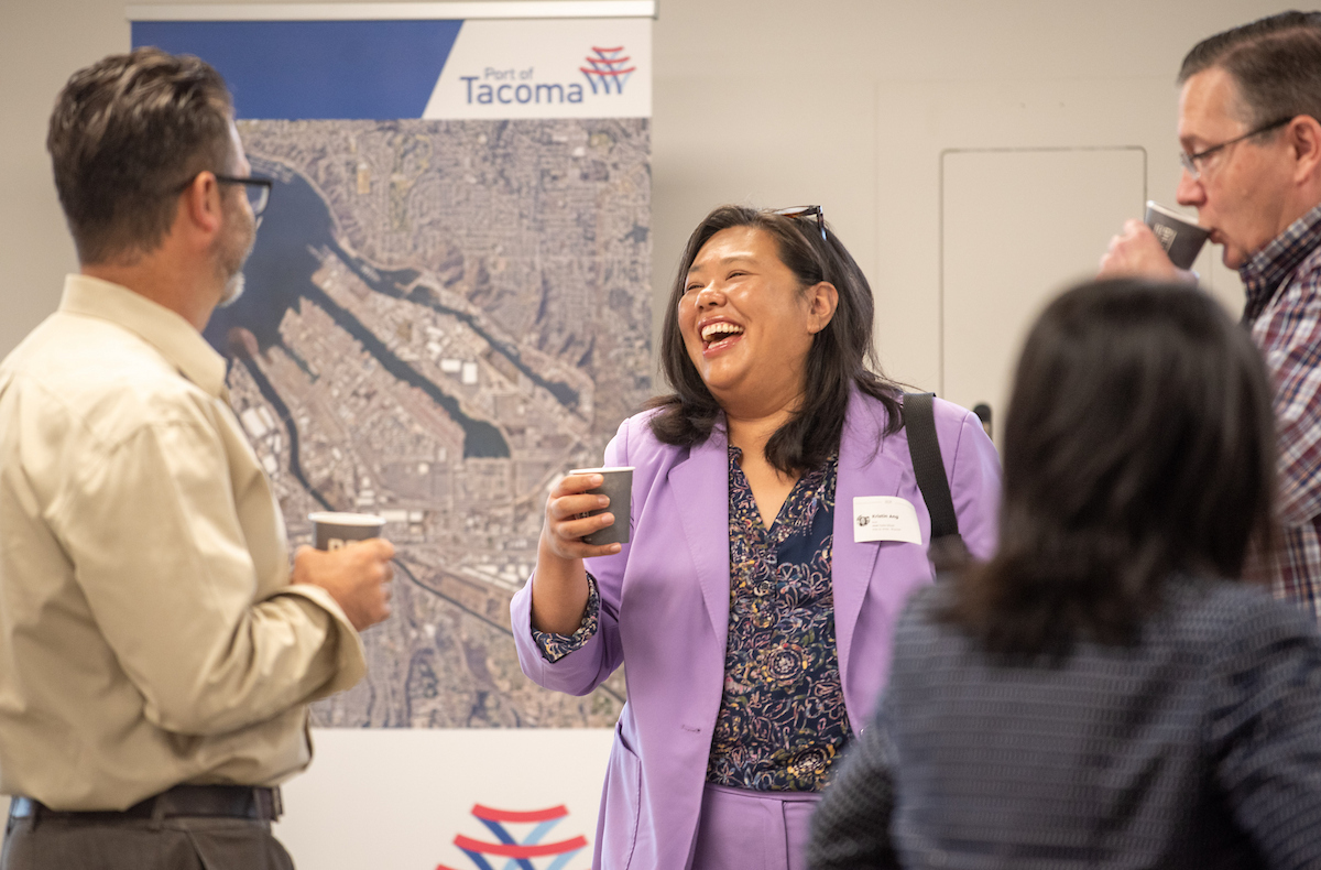 A woman in a lilac suit laughs while talking to a group of people drinking coffee.