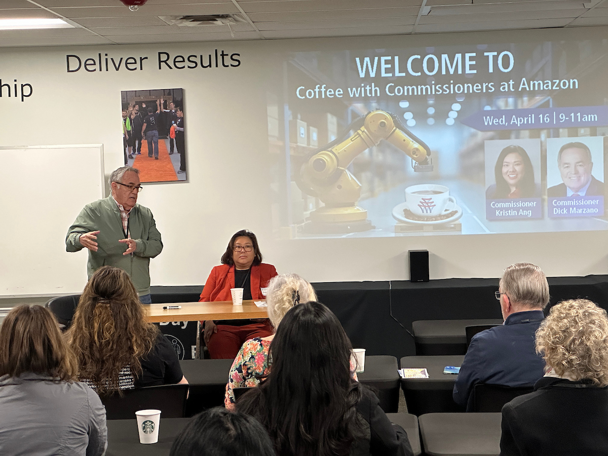 A man stands up and talks in front of a screen to a group of listeners in an office.