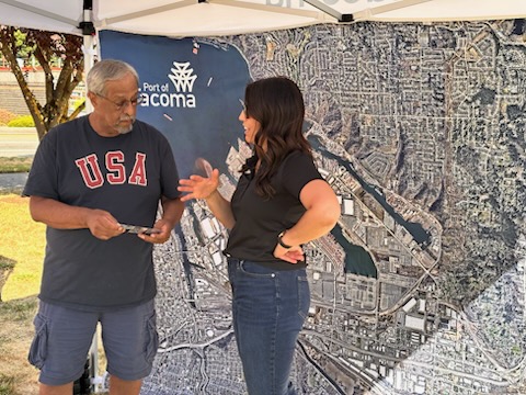 A woman and man standing in front of a large map speak to each other under a tent. 