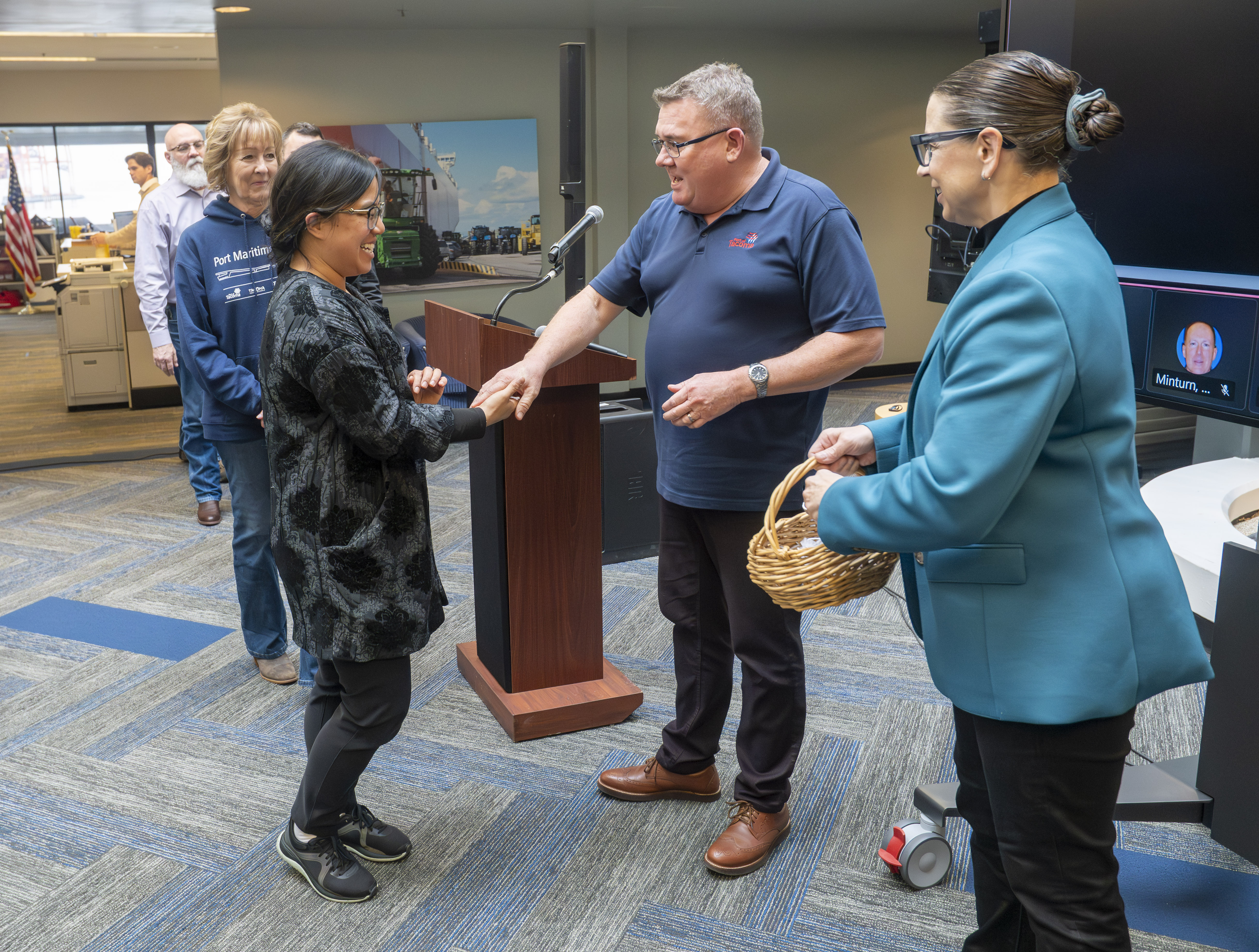 A man and woman shake hands in front of a podium with another woman holding a basket